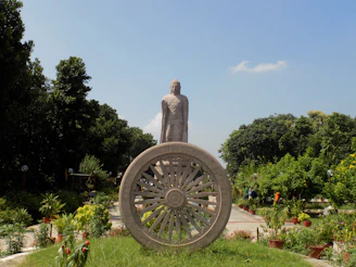 A large stone statue stands prominently in a garden-like area, surrounded by lush greenery and a clear blue sky. In the foreground, a decorative stone wheel is featured, adding a sense of historical or cultural significance.