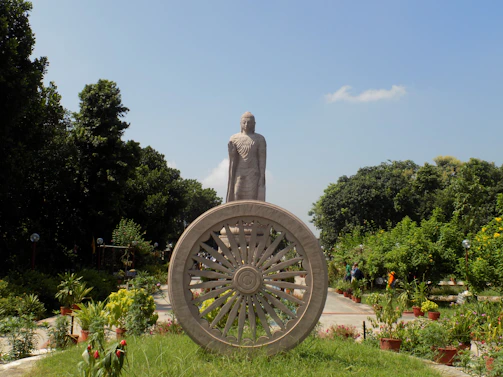 A large stone statue stands prominently in a garden-like area, surrounded by lush greenery and a clear blue sky. In the foreground, a decorative stone wheel is featured, adding a sense of historical or cultural significance.