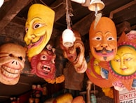 A collection of colorful, expressive masks hanging from a wooden structure. The masks vary in design, featuring large grins, mustaches, and exaggerated facial features. They are made of wood and painted in vibrant colors including yellow, red, and orange. There are also decorative elements like small flower garlands and motifs resembling the sun.