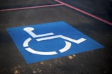 A blue square painted on an asphalt surface, featuring a white wheelchair symbol indicating a designated parking space for people with disabilities. Red lines are also visible on the pavement.