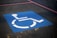 A blue square painted on an asphalt surface, featuring a white wheelchair symbol indicating a designated parking space for people with disabilities. Red lines are also visible on the pavement.