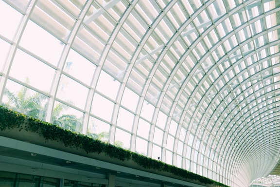 A modern architectural design featuring a large, curved glass roof with grid-like patterns. The ceiling allows natural light to stream in, illuminating the interior space. Lush green plants line the lower section of the glass panels, adding a touch of nature to the sleek, futuristic setting.