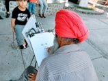 An artist with a red turban and striped shirt is seated and focused on sketching a portrait. In front of him, a young boy in a black shirt and grey pants is seated on a stool. The setting appears to be an outdoor area with stone and concrete surfaces.