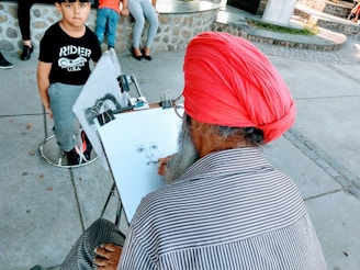 An artist with a red turban and striped shirt is seated and focused on sketching a portrait. In front of him, a young boy in a black shirt and grey pants is seated on a stool. The setting appears to be an outdoor area with stone and concrete surfaces.