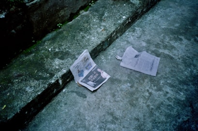 Two newspapers lie on a worn concrete step, one open displaying pages with text and images, and the other partly folded with visible text in a different language. The surrounding concrete appears aged with patches of green moss in the crevices.