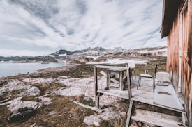 A rustic wooden cabin is situated on a rocky terrain near a calm body of water. The cabin has a small outdoor deck with simple wooden furniture, including a table and a couple of chairs. The landscape features snow-capped mountains in the distance and cloudy sky overhead, giving a sense of isolation and tranquility.