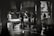 A black and white scene depicting several workers in activity, possibly in a market setting. The workers are standing around tables filled with ice, engaging in what appears to be the preparation or selling of goods. The environment is under a sheltered area with pillars, and the ground appears wet, reflecting the surrounding structures.