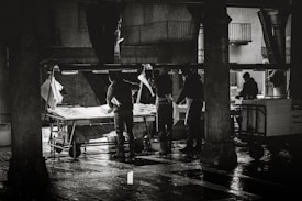 A black and white scene depicting several workers in activity, possibly in a market setting. The workers are standing around tables filled with ice, engaging in what appears to be the preparation or selling of goods. The environment is under a sheltered area with pillars, and the ground appears wet, reflecting the surrounding structures.