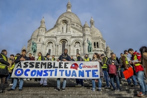 A group of people, many wearing yellow vests, gathers on the steps in front of a large, ornate building with a dome. They hold a banner with French text and a gear symbol, suggesting a political or social demonstration.