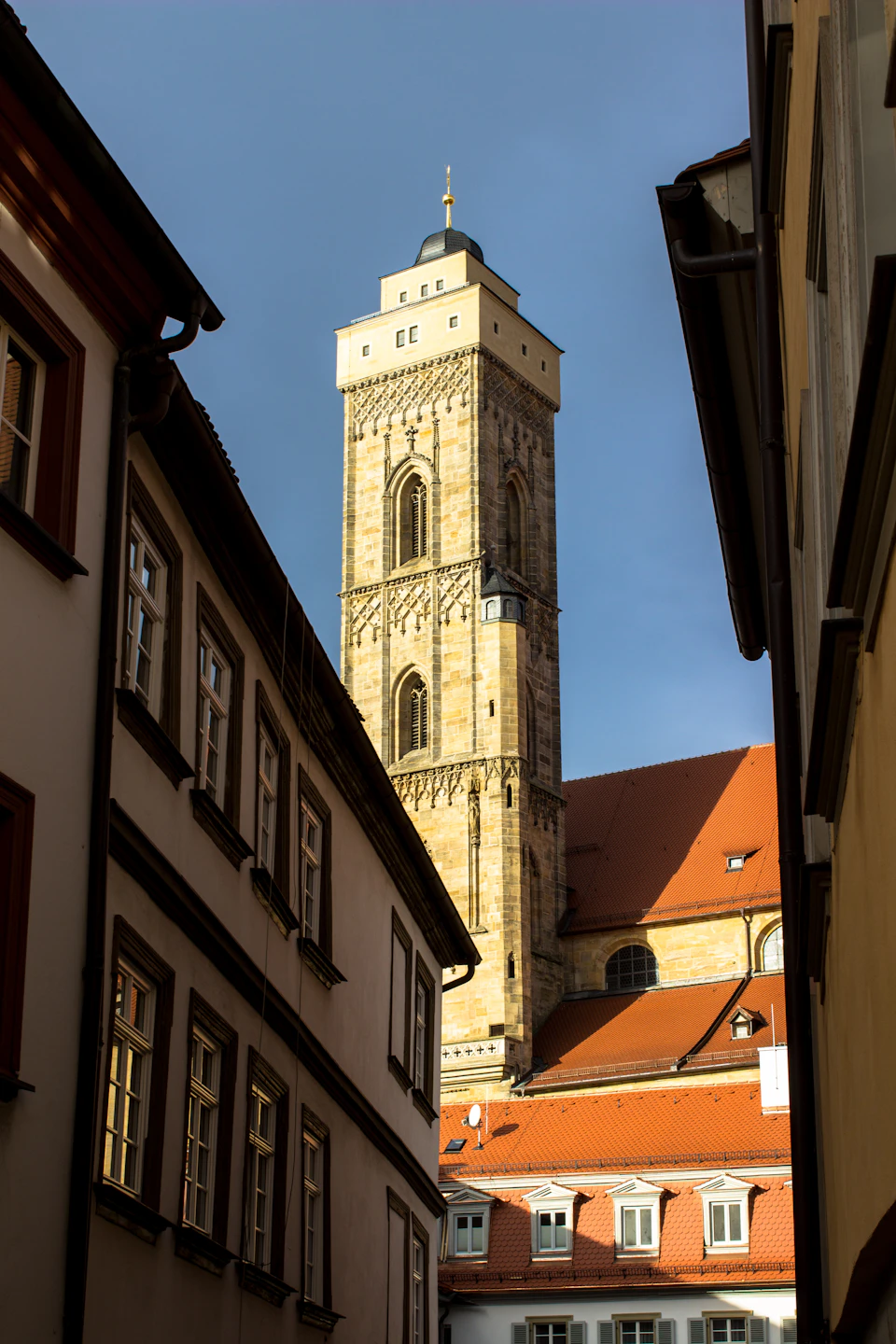 Tall stone building with clock tower stands beside a red-roofed building in European city street