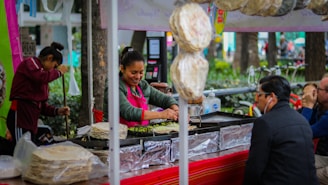 A street vendor with a cheerful expression is preparing food at an outdoor stall. Surrounding the vendor are stacks of tortillas, a grill, and various cooking utensils. Customers sit at the counter, engaging in conversation, while bicycles and a park-like setting are visible in the background.