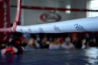 A close-up of a boxing ring featuring the white ropes with branding and a blurred background of spectators.