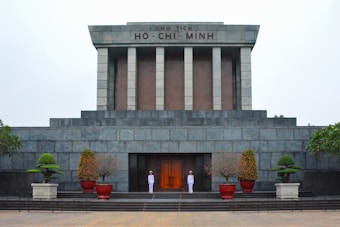 A large, solemn stone building with an inscription 'CHU TICH HO-CHI-MINH' at the top. Two uniformed guards stand at attention near the entrance. The structure features pillars and is surrounded by neatly trimmed plants in pots.