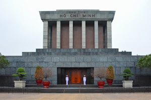 A large, solemn stone building with an inscription 'CHU TICH HO-CHI-MINH' at the top. Two uniformed guards stand at attention near the entrance. The structure features pillars and is surrounded by neatly trimmed plants in pots.