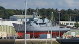 A large gray and red ship is positioned on land in a shipyard. It is surrounded by industrial buildings and green trees in the background. Several poles and masts are visible in the foreground, and boxes or containers are scattered around the base of the ship.