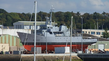 A large gray and red ship is positioned on land in a shipyard. It is surrounded by industrial buildings and green trees in the background. Several poles and masts are visible in the foreground, and boxes or containers are scattered around the base of the ship.