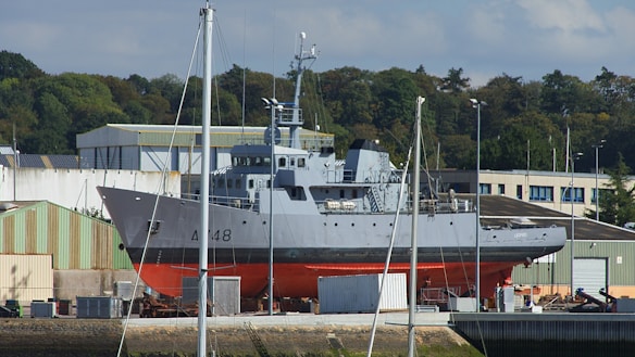 A large gray and red ship is positioned on land in a shipyard. It is surrounded by industrial buildings and green trees in the background. Several poles and masts are visible in the foreground, and boxes or containers are scattered around the base of the ship.