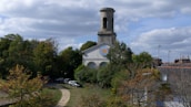 A historic church is surrounded by lush greenery with a tall, cylindrical tower rising above the trees. A small path leads up to the building, passing several parked cars. The sky is partly cloudy, casting soft shadows over the scene. Nearby rooftops are visible, adding a sense of a quaint, peaceful community.