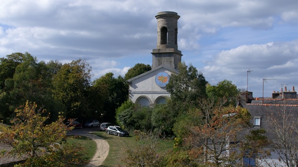 A historic church is surrounded by lush greenery with a tall, cylindrical tower rising above the trees. A small path leads up to the building, passing several parked cars. The sky is partly cloudy, casting soft shadows over the scene. Nearby rooftops are visible, adding a sense of a quaint, peaceful community.