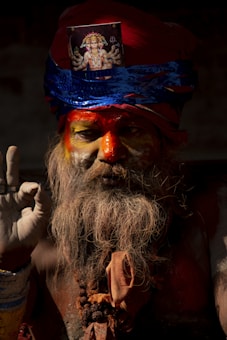 A person with a long beard and painted face poses while holding a hand in a gesture resembling a mudra. The figure wears a red and blue turban adorned with a religious image above the forehead. Rudraksha beads are around the neck, and the light casts shadows, creating a dramatic effect.