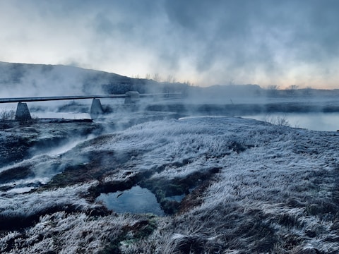 A frosty landscape with grass covered in ice and a few patches of water. Mist rises from the ground, suggesting geothermal activity. A bridge or pipeline structure extends across the scene, while the sky is overcast with hints of blue and orange from the setting or rising sun.