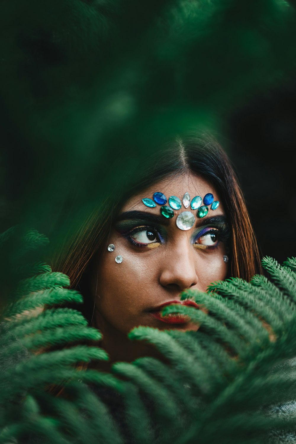 selective focus photography of woman with beads on forehead behind fern plant