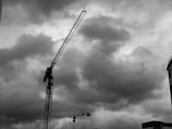 A black and white photograph of a construction site with two large cranes towering against a backdrop of dramatic, cloudy skies. The image captures the cranes in the foreground, with their long arms extending upwards. The silhouette of a building is visible in the bottom right corner, enhancing the urban setting.