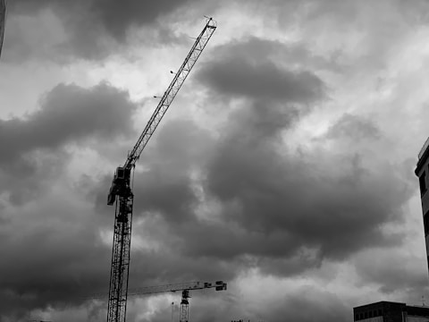A black and white photograph of a construction site with two large cranes towering against a backdrop of dramatic, cloudy skies. The image captures the cranes in the foreground, with their long arms extending upwards. The silhouette of a building is visible in the bottom right corner, enhancing the urban setting.