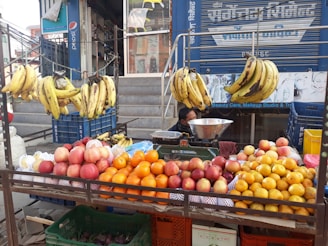 A street vendor's fruit stand displays a variety of fruits including bananas hanging from a rack, and piles of apples, oranges, and other citrus fruits arranged on a table. The vendor is partially visible, sitting behind the display with a metal bowl and weighing scale. The background features a storefront with signs and advertisements.