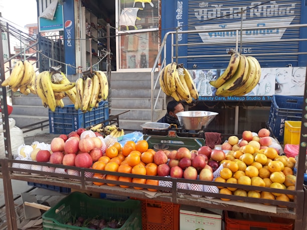 A street vendor's fruit stand displays a variety of fruits including bananas hanging from a rack, and piles of apples, oranges, and other citrus fruits arranged on a table. The vendor is partially visible, sitting behind the display with a metal bowl and weighing scale. The background features a storefront with signs and advertisements.