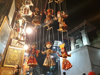 Several traditional string puppets hang against a nighttime backdrop, illuminated by warm lights. The puppets display intricate costumes with various vibrant colors, showcasing cultural artistry. A large bell and architectural structures are visible in the background.