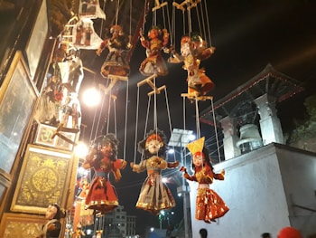 Several traditional string puppets hang against a nighttime backdrop, illuminated by warm lights. The puppets display intricate costumes with various vibrant colors, showcasing cultural artistry. A large bell and architectural structures are visible in the background.