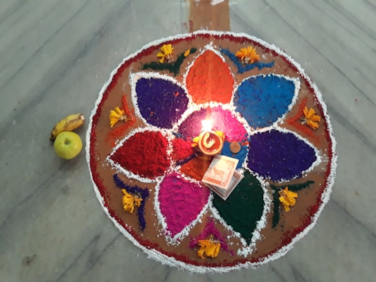 A colorful rangoli design on a marble floor features six large petal shapes filled with vibrant colors such as purple, blue, pink, red, and green. The design is accentuated with yellow flowers placed around it. At the center, there is a lit oil lamp surrounded by a coin, a small booklet, and an incense stick packet. To the side, a banana and an apple are placed next to the rangoli.