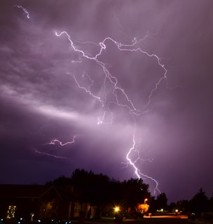 Dark night sky with multiple lightning bolts illuminating the clouds and striking the ground. The image shows a residential area with houses and trees. Streetlights and outdoor lights create small, warm glows among the dark surroundings. The lightning branches out in various directions, creating a dramatic visual effect.
