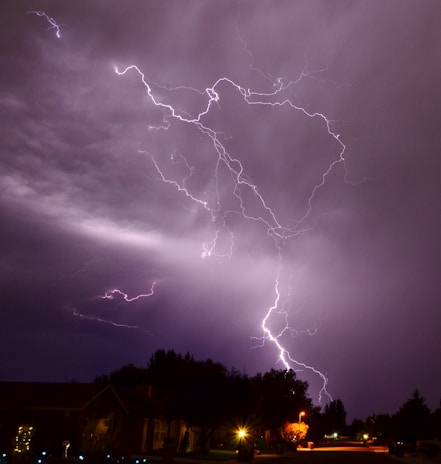 Dark night sky with multiple lightning bolts illuminating the clouds and striking the ground. The image shows a residential area with houses and trees. Streetlights and outdoor lights create small, warm glows among the dark surroundings. The lightning branches out in various directions, creating a dramatic visual effect.