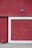 A large red wooden wall featuring a section with diagonal planks framed by a white border. Above, a black industrial-style light fixture is mounted, adding a touch of rustic charm. The ground is made of concrete with some grass visible at the edge.