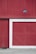 A large red wooden wall featuring a section with diagonal planks framed by a white border. Above, a black industrial-style light fixture is mounted, adding a touch of rustic charm. The ground is made of concrete with some grass visible at the edge.