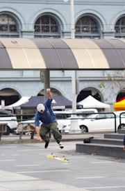 A person in motion performing a skateboard trick in an urban setting. The skateboarder is airborne, wearing a blue shirt, olive-green pants, and a white cap. The background features a building with arched windows and a symmetrical design. There are also parked cars and market tents visible in the street scene.