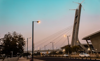 A modern urban scene featuring a tall structure with a wind turbine on top. Several cables extend from the structure, resembling a suspension bridge. Below the structure, there are street lamps illuminating a road where a few cars are visible. On the left, there is a tree and some buildings in the background. The sky has a gradient ranging from blue to a light pink hue.