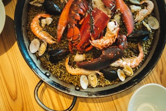 A close-up of a seafood dish featuring a large lobster, shrimp, clams, mussels, and other shellfish arranged on a bed of rice in a round, shallow pan resting on a wooden table.