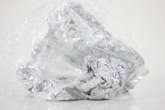 Crumbled foil loosely wrapped in a plastic sheet on a plain background. The plastic is transparent and the foil is shiny with a metallic luster.