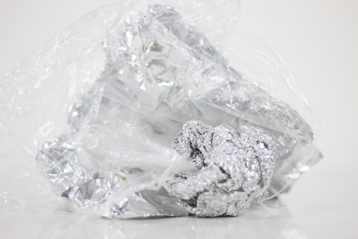 Crumbled foil loosely wrapped in a plastic sheet on a plain background. The plastic is transparent and the foil is shiny with a metallic luster.