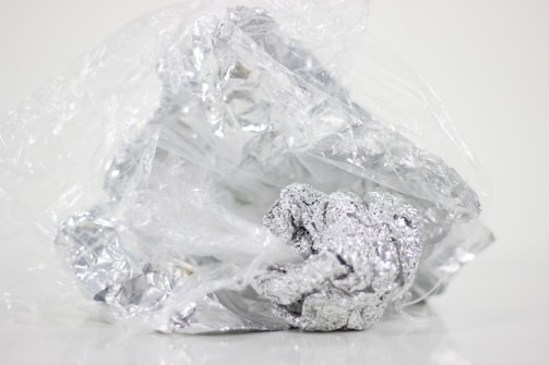 Crumbled foil loosely wrapped in a plastic sheet on a plain background. The plastic is transparent and the foil is shiny with a metallic luster.