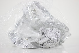 Crumbled foil loosely wrapped in a plastic sheet on a plain background. The plastic is transparent and the foil is shiny with a metallic luster.
