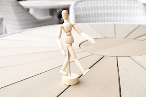 A wooden mannequin posed in a walking stance is positioned on a round, light-colored wooden table. The background features soft-focus, neutral-colored woven chairs.