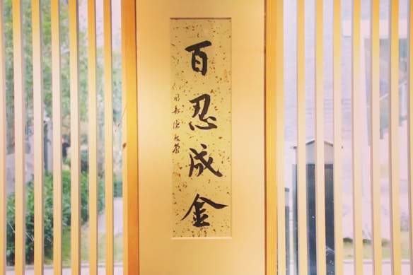 A vertical banner with Asian calligraphy hangs between wooden blinds. Light filters through, casting soft shadows inside, while greenery is faintly visible outside.