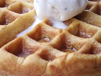 A close-up view of a golden-brown waffle topped with a scoop of melting vanilla ice cream. The texture of the waffle is crisp, with visible pockets of syrup glistening in the light.