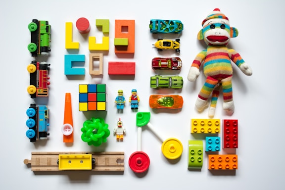 A collection of colorful toys arranged on a white surface, including wooden blocks, a sock monkey, toy cars, a Rubik's cube, train pieces, LEGO bricks, a green ring, a miniature traffic cone, a lollipop toy, and small figurines.
