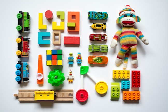 A collection of colorful toys arranged on a white surface, including wooden blocks, a sock monkey, toy cars, a Rubik's cube, train pieces, LEGO bricks, a green ring, a miniature traffic cone, a lollipop toy, and small figurines.