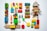 A collection of colorful toys arranged on a white surface, including wooden blocks, a sock monkey, toy cars, a Rubik's cube, train pieces, LEGO bricks, a green ring, a miniature traffic cone, a lollipop toy, and small figurines.
